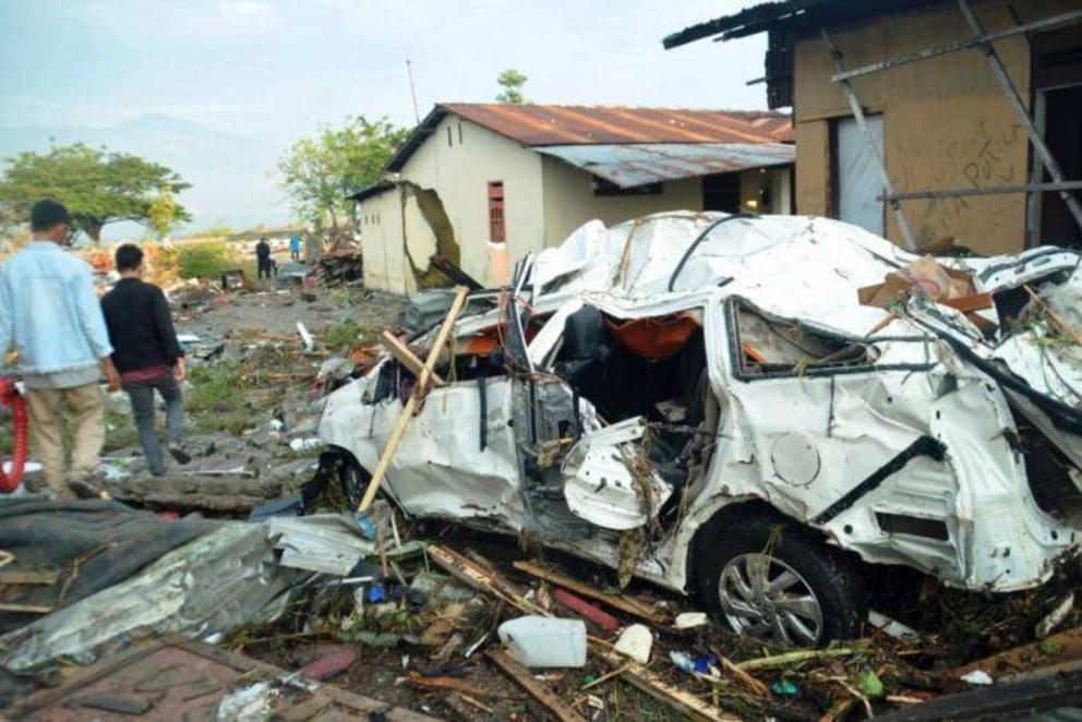 Indonesian men walk past the wreckage of a car.